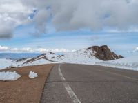 an empty road with snow and mountains in the back ground under cloudy skies on a sunny day