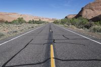 the empty road that is running through the desert land and mountains in the background with the shadows on the pavement