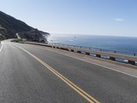 a car drives down a curvy road along the ocean side with some hills in the background