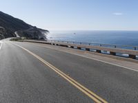 a car drives down a curvy road along the ocean side with some hills in the background