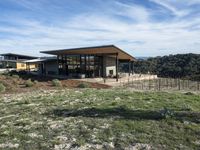 a house with a wooden roof stands on some grassy land next to a fence and mountain range