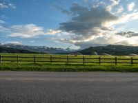 the road is empty during the day and has mountains in the distance as well as a fence