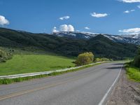 the empty road is going through a valley with mountains and grass in the background and two houses on both sides