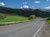 the empty road is going through a valley with mountains and grass in the background and two houses on both sides