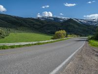 the empty road is going through a valley with mountains and grass in the background and two houses on both sides