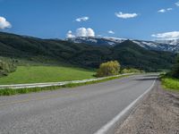 the empty road is going through a valley with mountains and grass in the background and two houses on both sides