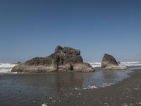 two rocks with one rock sticking out into the water, sitting on a black sand beach