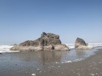 two rocks with one rock sticking out into the water, sitting on a black sand beach