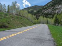 road in the mountains with grass and trees and a fence surrounding it on a bright day