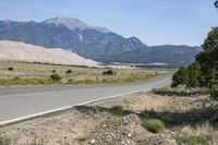 a road winds away from a desert hill in the background, with mountains and trees on both sides