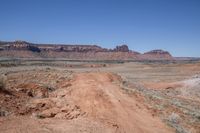 a dirt road through a desert plain with a mountain behind it and a clear blue sky in the background