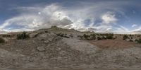 a big dirt hill in the desert under a cloudy blue sky and white clouds with one large red spot in front