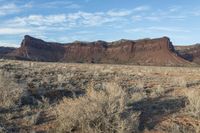 a hill with some brown dirt and sparse shrubs in front of it under a partly cloudy blue sky