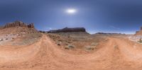 a picture of dirt road going through desert land with rock cliff in background and sun shining