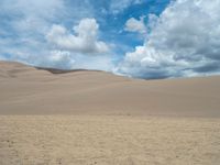 the man is riding his horse through the large expanse of sand dunes with blue sky and clouds over them