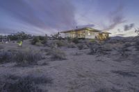 the house is on an arid plain covered by brush and bushes at dusk, near some light poles