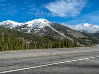 a snowy mountainside with a blue sky with some clouds in it, and trees, on the right side of the highway