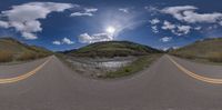 a fisheye picture shows the sun in a cloudy sky over an empty road with mountains