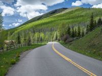 road in the mountains with grass and trees and a fence surrounding it on a bright day