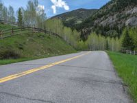 road in the mountains with grass and trees and a fence surrounding it on a bright day