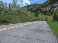 road in the mountains with grass and trees and a fence surrounding it on a bright day