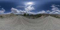 a panoramic picture shows the sun in between a dirt road and hills and clouds in the sky