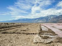 a body of water next to a hill covered in dirt and mud and mountains in the background