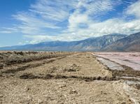 a body of water next to a hill covered in dirt and mud and mountains in the background