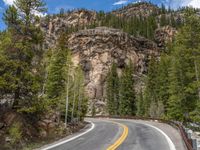 the curved road curves in front of a mountain area with pine trees behind it,