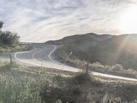 the sun shines brightly on a curved road that runs past a hill and is bordered by grass and trees