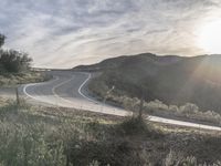 the sun shines brightly on a curved road that runs past a hill and is bordered by grass and trees