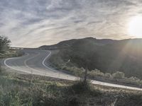 the sun shines brightly on a curved road that runs past a hill and is bordered by grass and trees