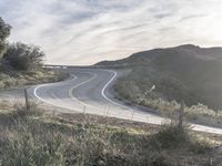 the sun shines brightly on a curved road that runs past a hill and is bordered by grass and trees