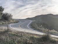 the sun shines brightly on a curved road that runs past a hill and is bordered by grass and trees