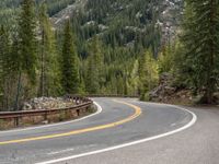 the curved road curves in front of a mountain area with pine trees behind it,