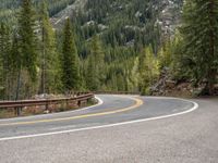 the curved road curves in front of a mountain area with pine trees behind it,