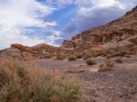 a dirt field surrounded by rocks under a cloudy sky in the desert with shrubs and shrubs around it