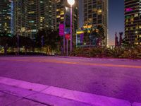 an empty street at night with street lights lit up in front of skyscrapers and a pink street light