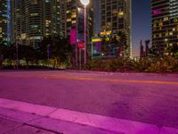 an empty street at night with street lights lit up in front of skyscrapers and a pink street light