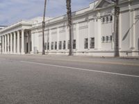 a white building sitting on the side of a road with a few tall palm trees