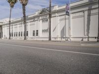 a white building sitting on the side of a road with a few tall palm trees