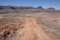 a dirt road through a desert plain with a mountain behind it and a clear blue sky in the background