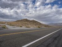 the empty road is lined with yellow lines and grey clouds and mountains in the distance