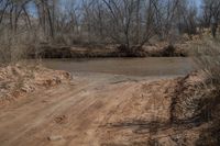 a dirty, dirt road with a river behind it and trees around the intersection behind