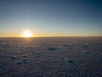 a field of snow with the sun setting on top of it in a background of some horizon land