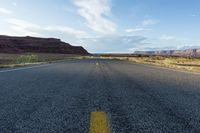 an empty road with yellow lines, dirt and mountains in the distance and a blue sky