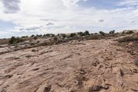 a dirt area with a very cloudy sky above it and bushes in the background and rocks and shrubs on each side