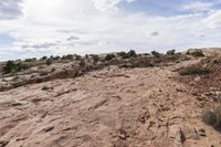 a dirt area with a very cloudy sky above it and bushes in the background and rocks and shrubs on each side