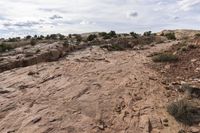 a dirt area with a very cloudy sky above it and bushes in the background and rocks and shrubs on each side