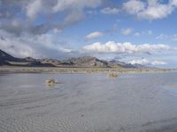 an empty field that is surrounded by some mountains in the distance with clouds overhead and sand on the ground